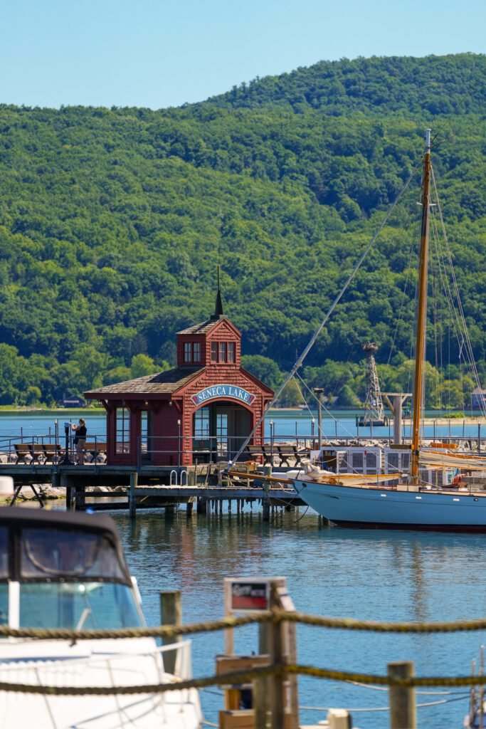 Red shingled boathouse with a Seneca Lake sign, a moored sailboat and forested hills across the water