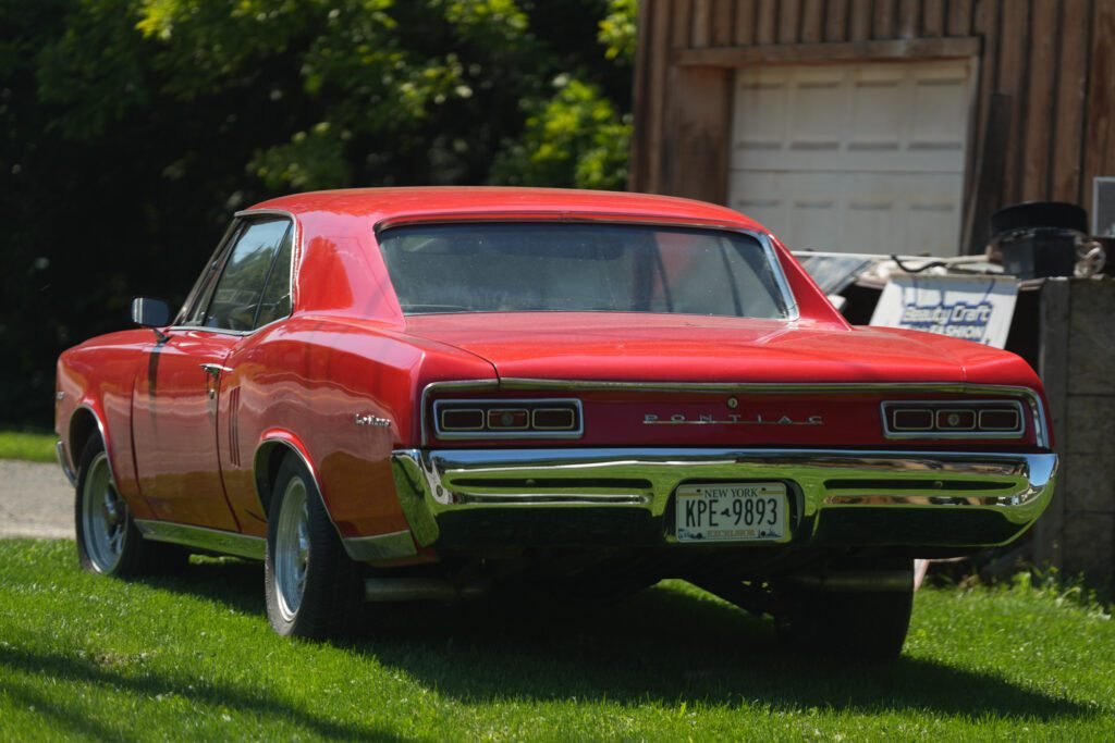 A red 1960s Pontiac parked on grass beside a weathered barn with New York plates