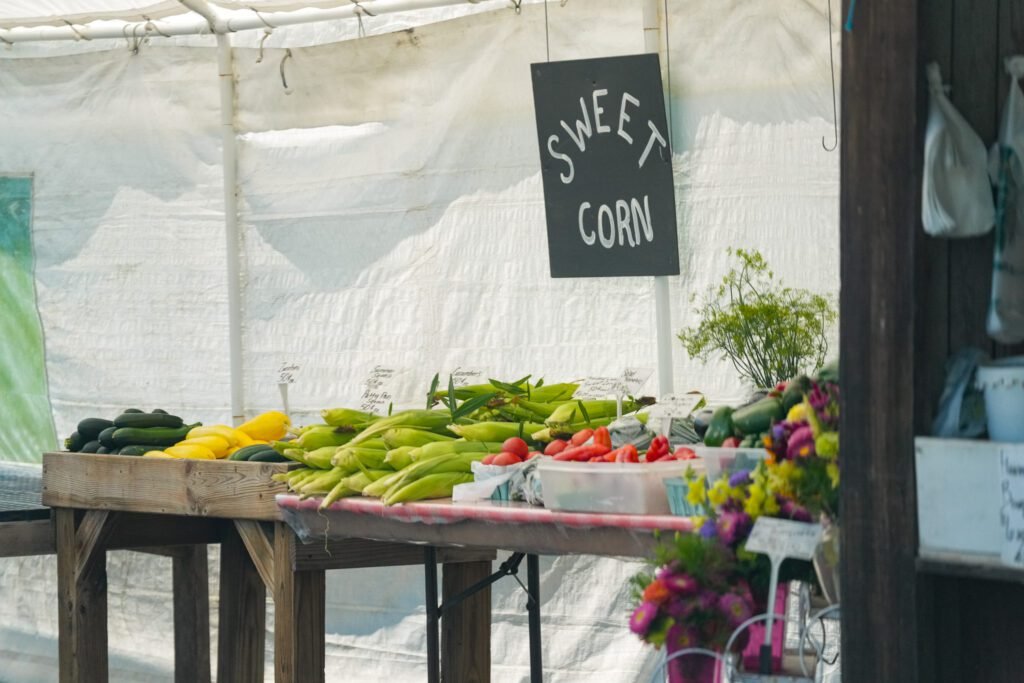 Sweet corn, yellow squash and peppers on a farm stand table beneath a hand-lettered Sweet Corn sign