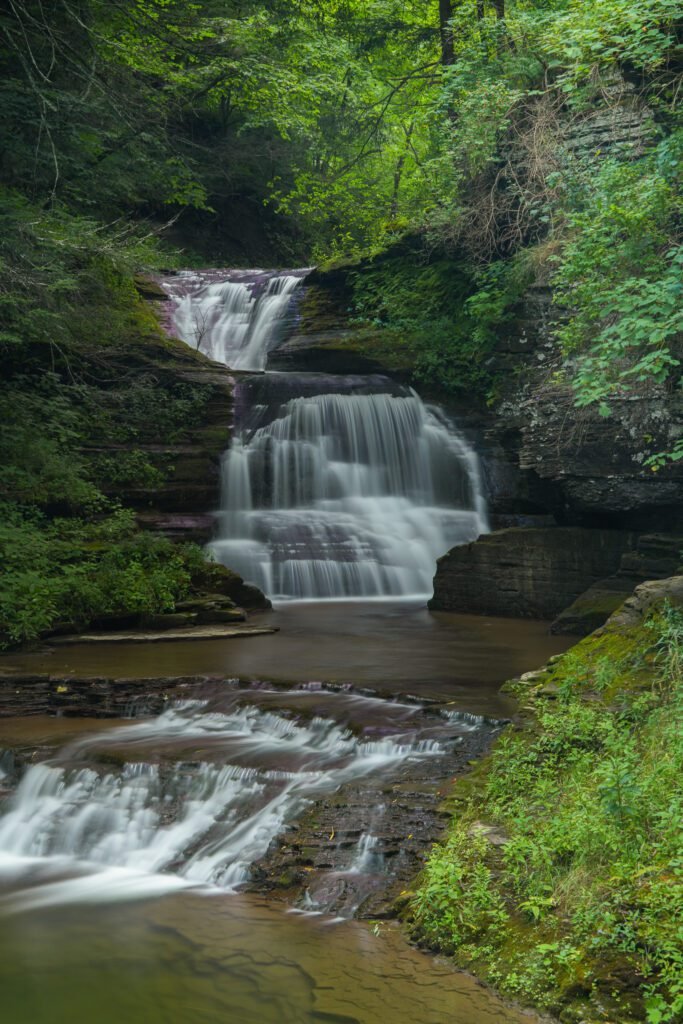 A tiered waterfall dropping over dark rock shelves into a still pool in a wooded gorge at Robert H. Treman State Park