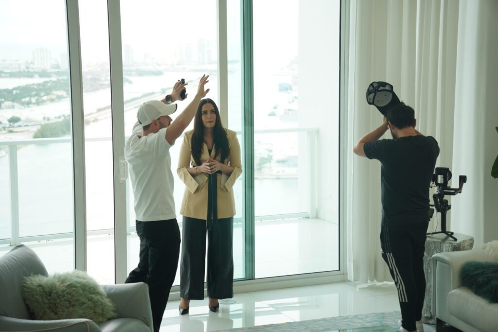 Behind the scenes on a Miami real estate video, framing Chava Feigen by the floor to ceiling windows with the bay behind her while a second operator sets up a gimbal