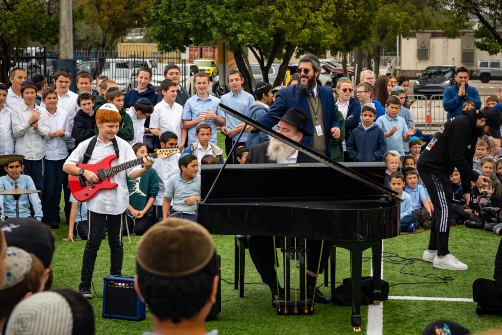 A student playing a red electric guitar beside a grand piano on the field as the school watches, Gift to the Rebbe music video