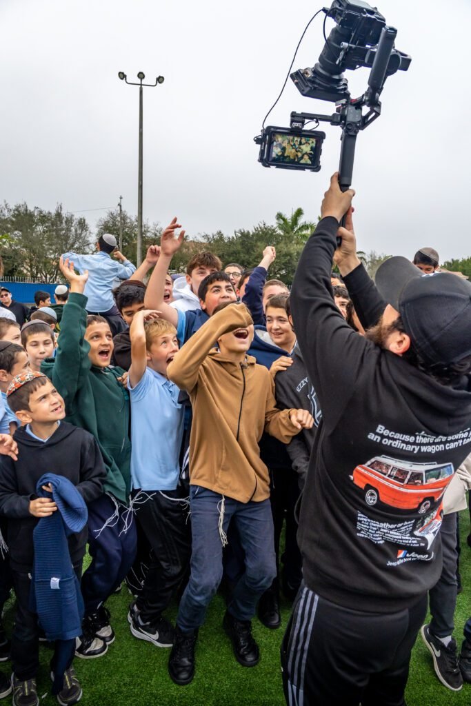 A camera operator holding a gimbal rig above a crowd of cheering students while filming the Gift to the Rebbe music video