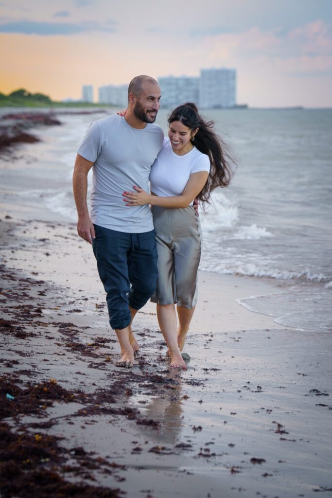 Couple walking barefoot at the water's edge under a pink and gold sky