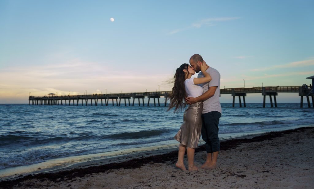 Couple embracing on the beach with a long pier and a rising moon behind them
