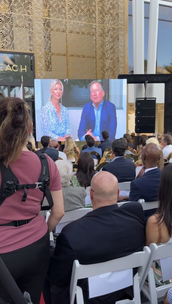 Outdoor audience watching a large screen showing two people seated in an interview