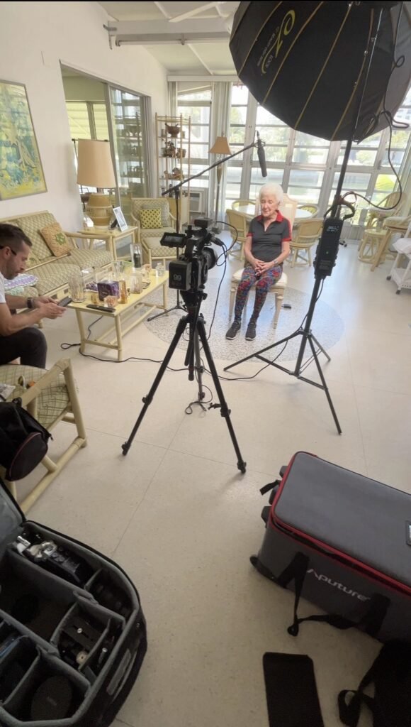 Older woman seated for an interview in a sunlit living room, boom microphone and softbox overhead