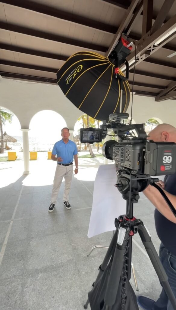 Tripod-mounted camera and overhead softbox pointed at a man in a blue polo under a beachfront pavilion