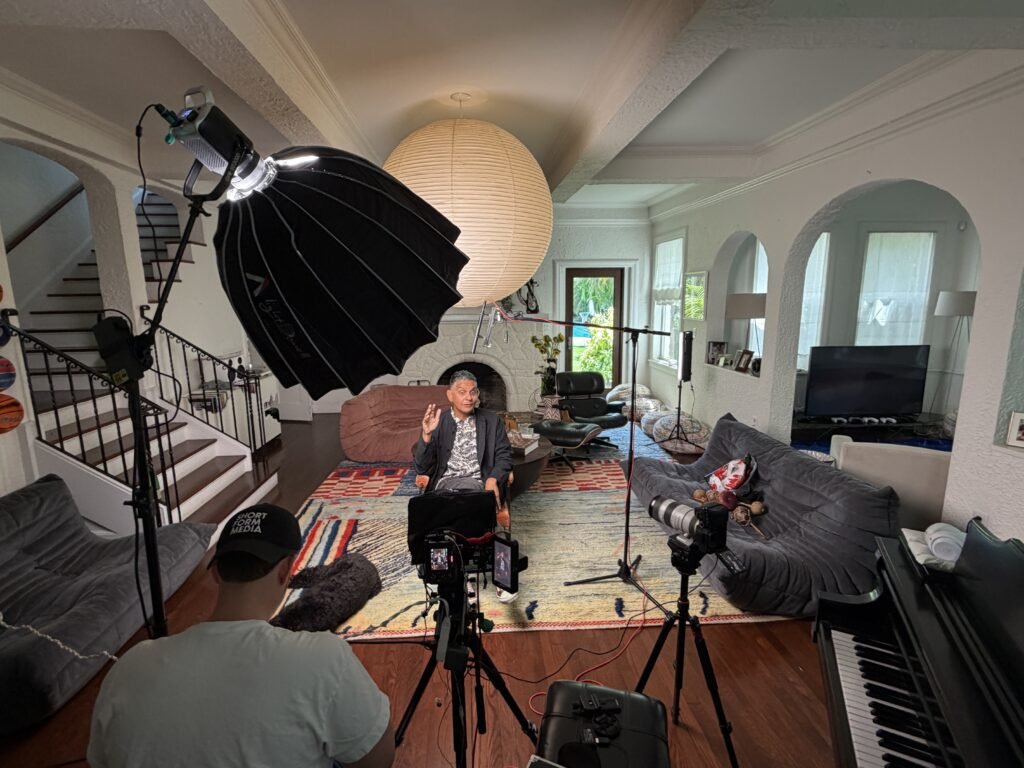 Two-camera interview set up in a living room for a Short Form Media production, the subject mid-sentence on a low sofa under a large softbox while a crew member in a Short Form Media cap operates in the foreground