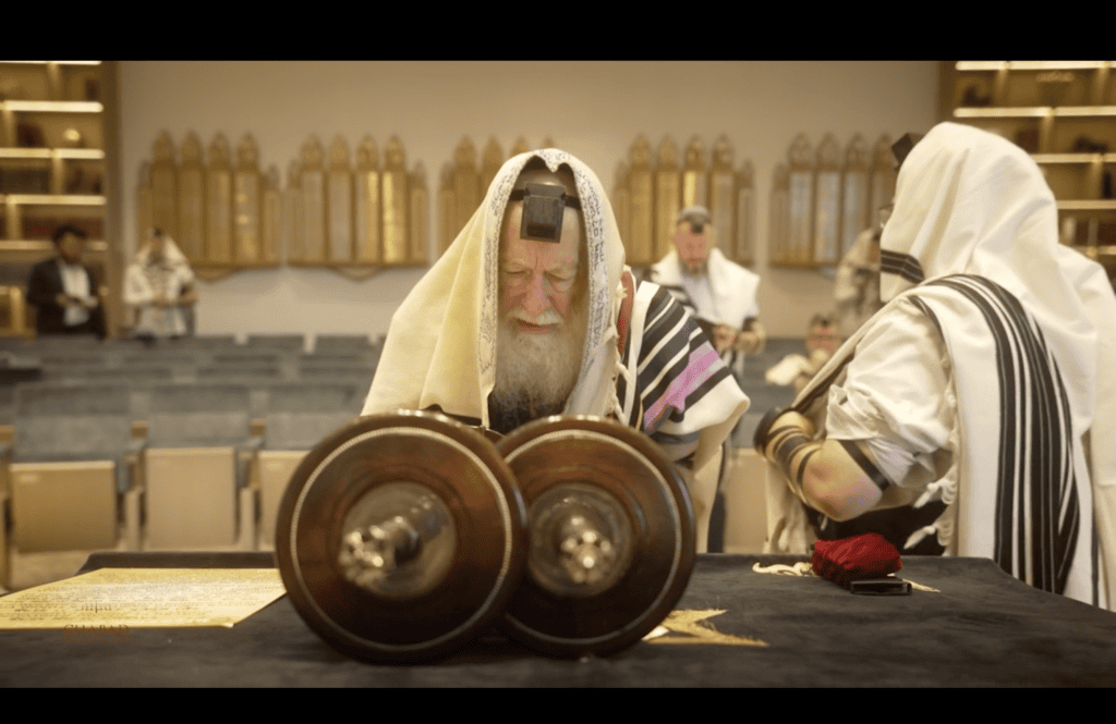 An elder in tallit and tefillin reading from an open Torah scroll on the bimah