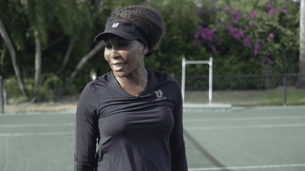Venus Williams on her home court in a white top, bougainvillea flowering behind the fence