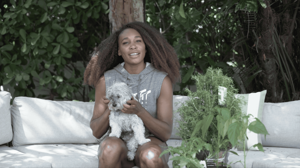 Venus Williams sitting on an outdoor sofa holding her small white dog, herb plants beside her