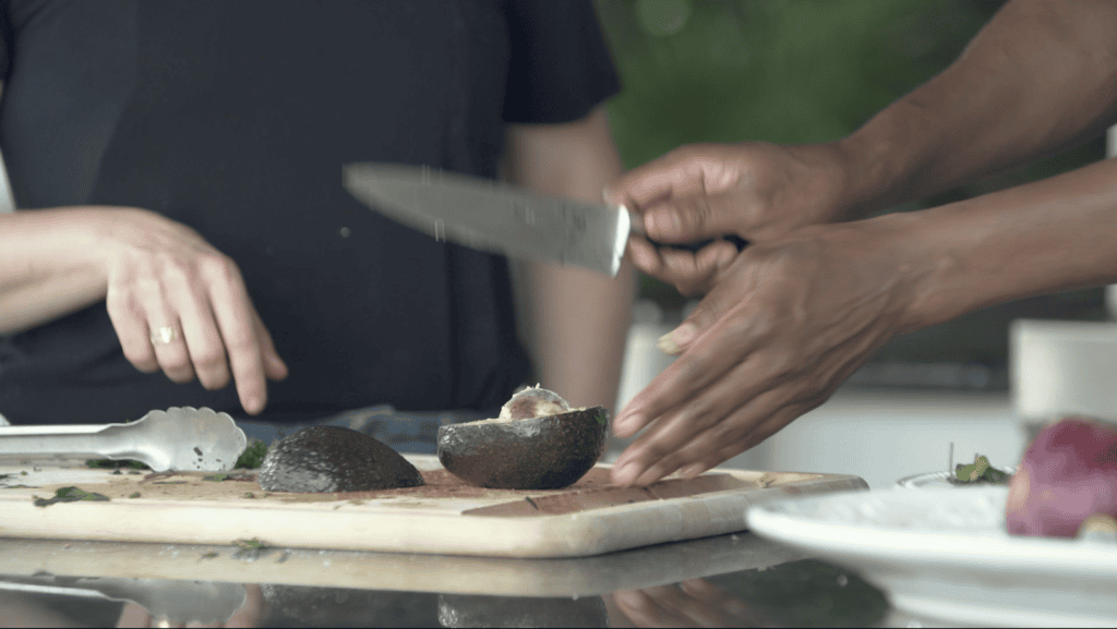 Two pairs of hands halving an avocado on a wooden board in an outdoor kitchen