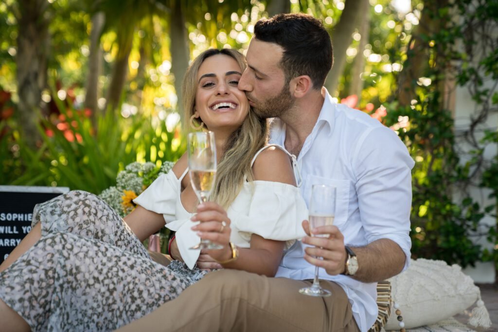 Man in white shirt kissing smiling blonde woman's cheek, both holding champagne flutes outdoors