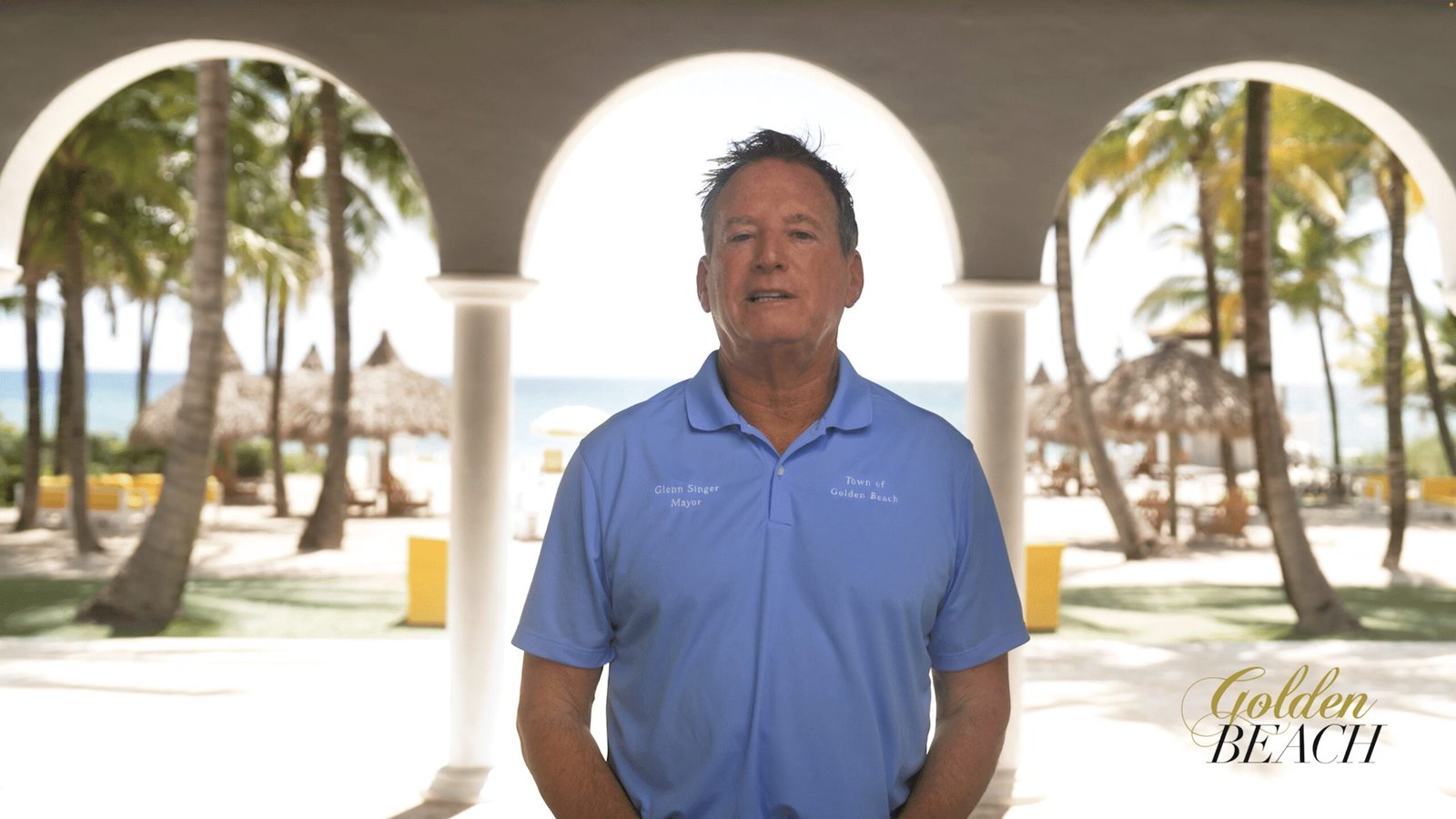 Mayor Glenn Singer speaking to camera under the arches of the Golden Beach Beach Pavilion, palms and ocean behind him