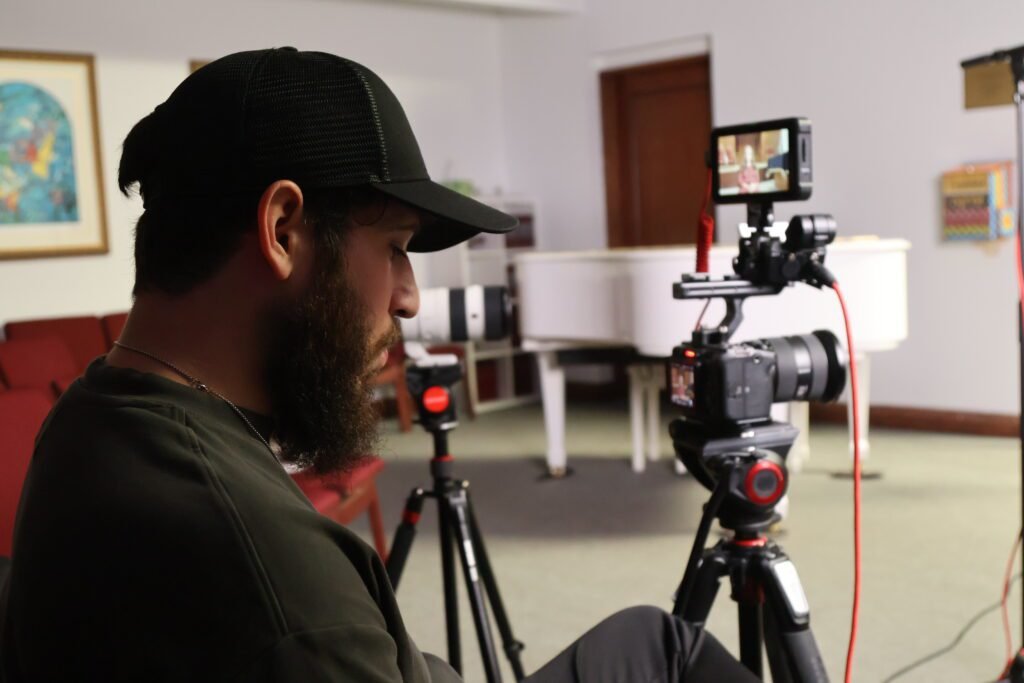Dov Gurewicz behind the camera in profile, the monitor framing the interview with a white grand piano behind him
