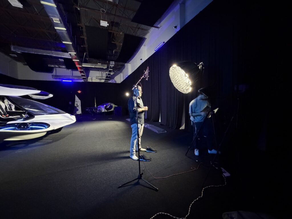 Wide of the darkened Doroni studio during filming, a presenter at the boom microphone stand with the round softbox lit and the nose of the H1-X at the edge of frame