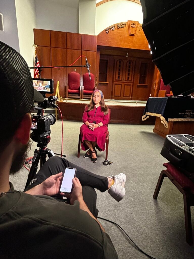 Interview set up in the Lanaar School sanctuary, a woman in a magenta dress seated before the ark while the camera, boom and softbox are framed from behind the operator