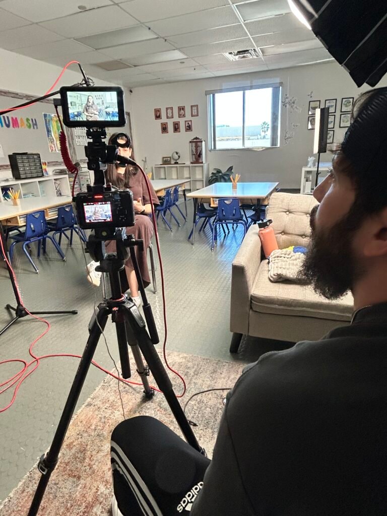 Filming a woman seated for an interview in a Lanaar School classroom, the camera monitor framing her while Dov Gurewicz watches from behind the rig