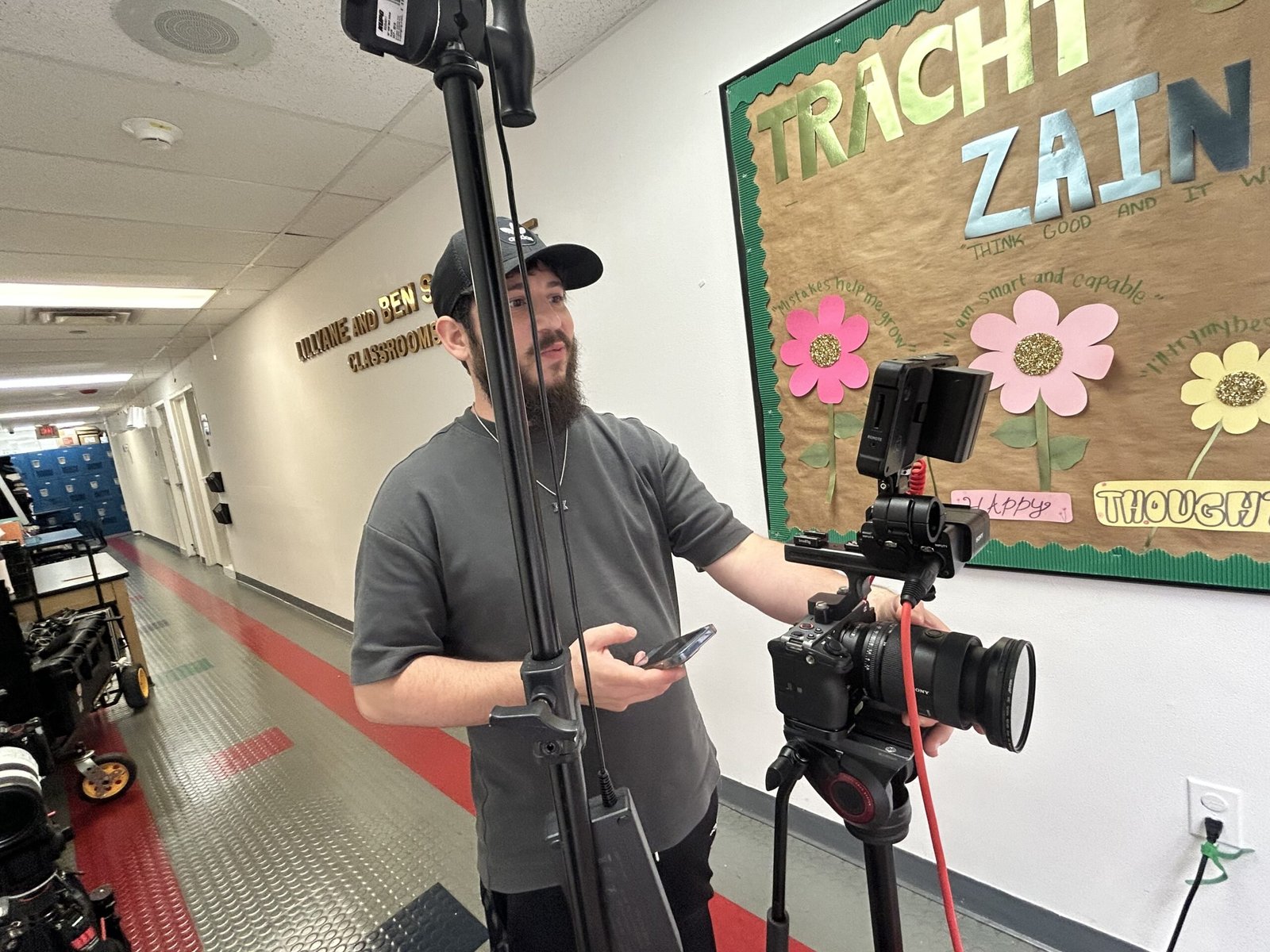 Dov Gurewicz moving a light stand and camera between rooms in the Lanaar School hallway, gear cases on the floor behind him